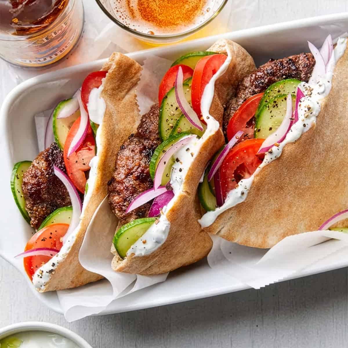 Three pita sandwiches filled with seasoned meat, sliced cucumbers, tomatoes, red onions, and a dollop of white sauce-an easy May one pot dinner-served in a tray with drinks visible in the background.