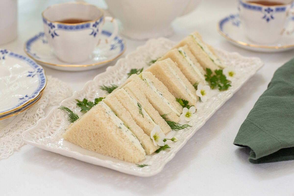A rectangular platter with six triangular Benedictine Tea Sandwiches, garnished with dill, parsley, and small white flowers, sits on a white tablecloth near teacups and a green napkin.