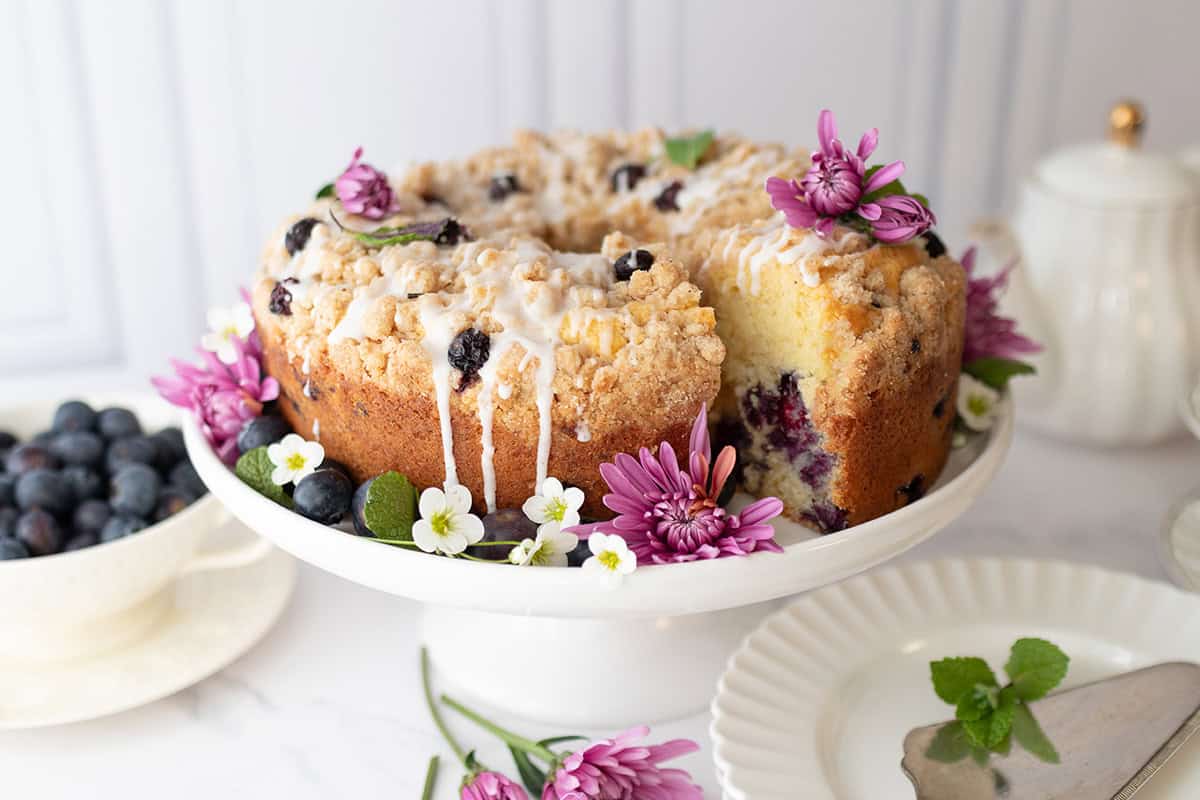 A blueberry yogurt teacake with crumb topping and icing sits on a white cake stand, decorated with purple flowers and surrounded by a plate, serving utensil, and bowl of blueberries.