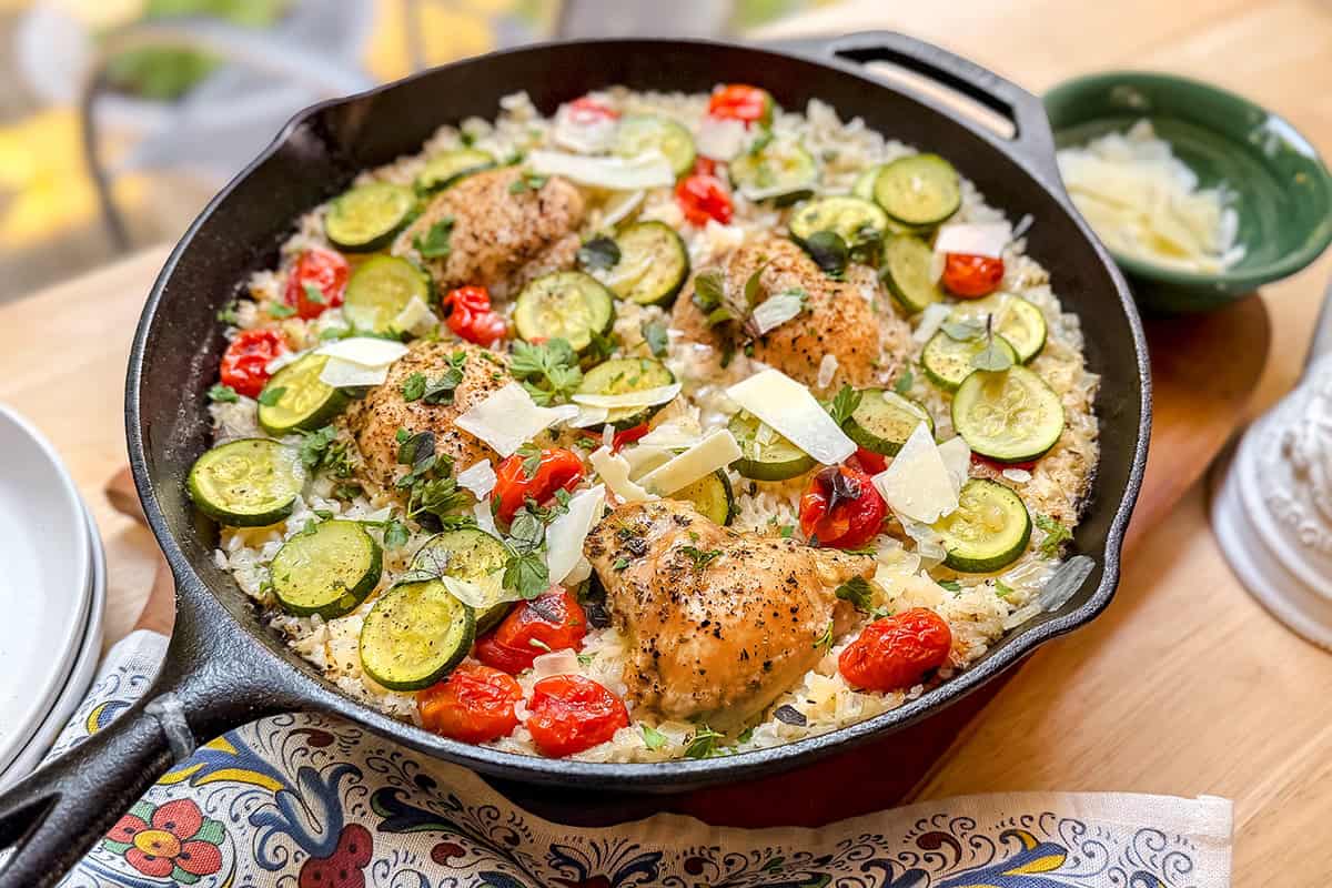 A one pan skillet filled with Tuscan chicken thighs, sliced zucchini, cherry tomatoes, rice, herbs, and shaved cheese rests on a wooden table.
