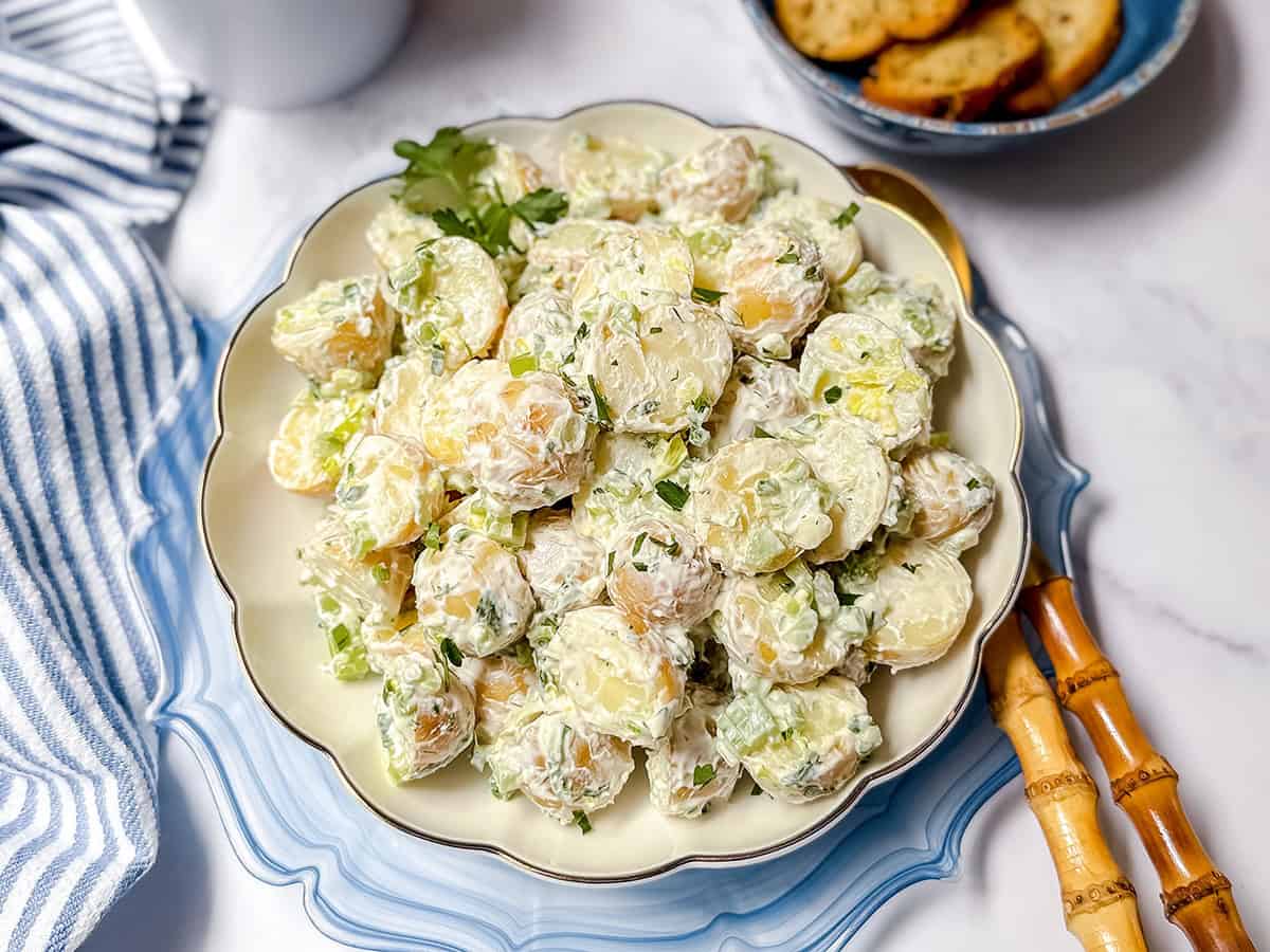 A plate of creamy Yukon Gold potato salad garnished with chopped herbs, placed on a scalloped dish next to a blue plate of toasted bread slices and bamboo-handled cutlery.