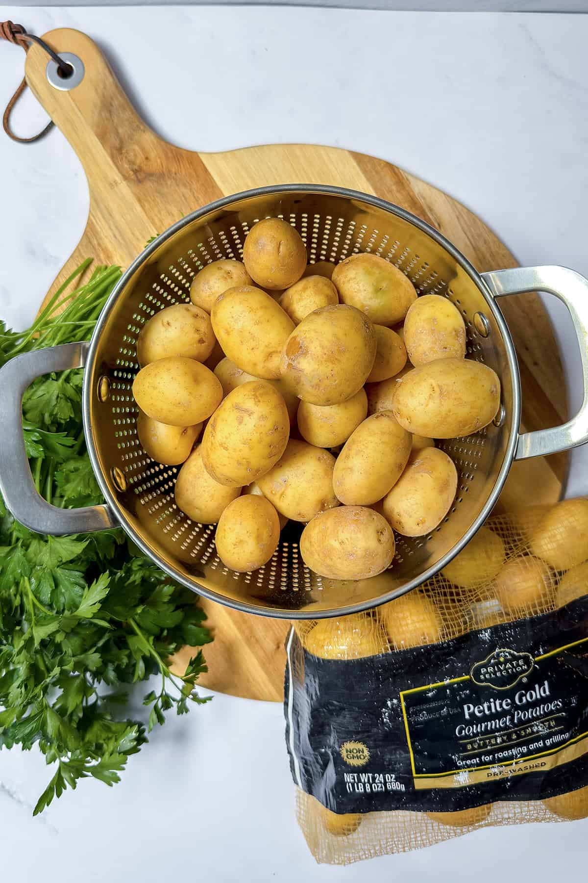 A metal colander filled with small gold potatoes sits on a wooden cutting board next to a bunch of parsley and a bag labeled "Petite Gold Gourmet Potatoes," perfect for making creamy Yukon Gold potato salad.