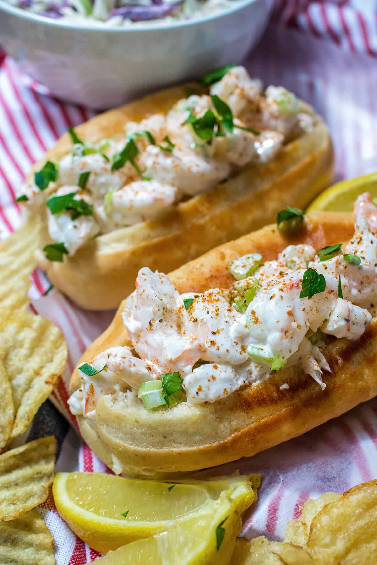 Two New England style shrimp rolls with creamy filling and herbs on toasted buns, garnished with paprika, served with potato chips and lemon wedges.
