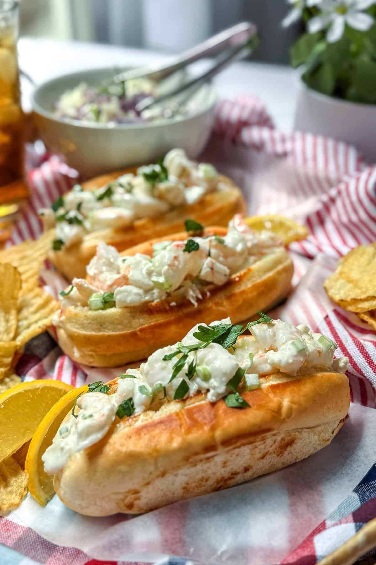 Three New England style shrimp rolls topped with chopped herbs are served on a striped cloth with potato chips and lemon wedges, with a bowl of coleslaw in the background.