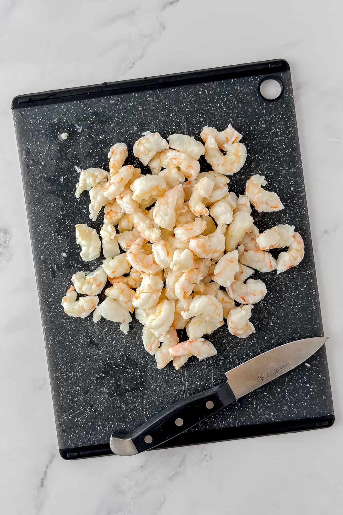 Peeled and deveined shrimp pieces on a black cutting board with a knife beside them, perfect for preparing classic New England seafood dishes like Shrimp Rolls.