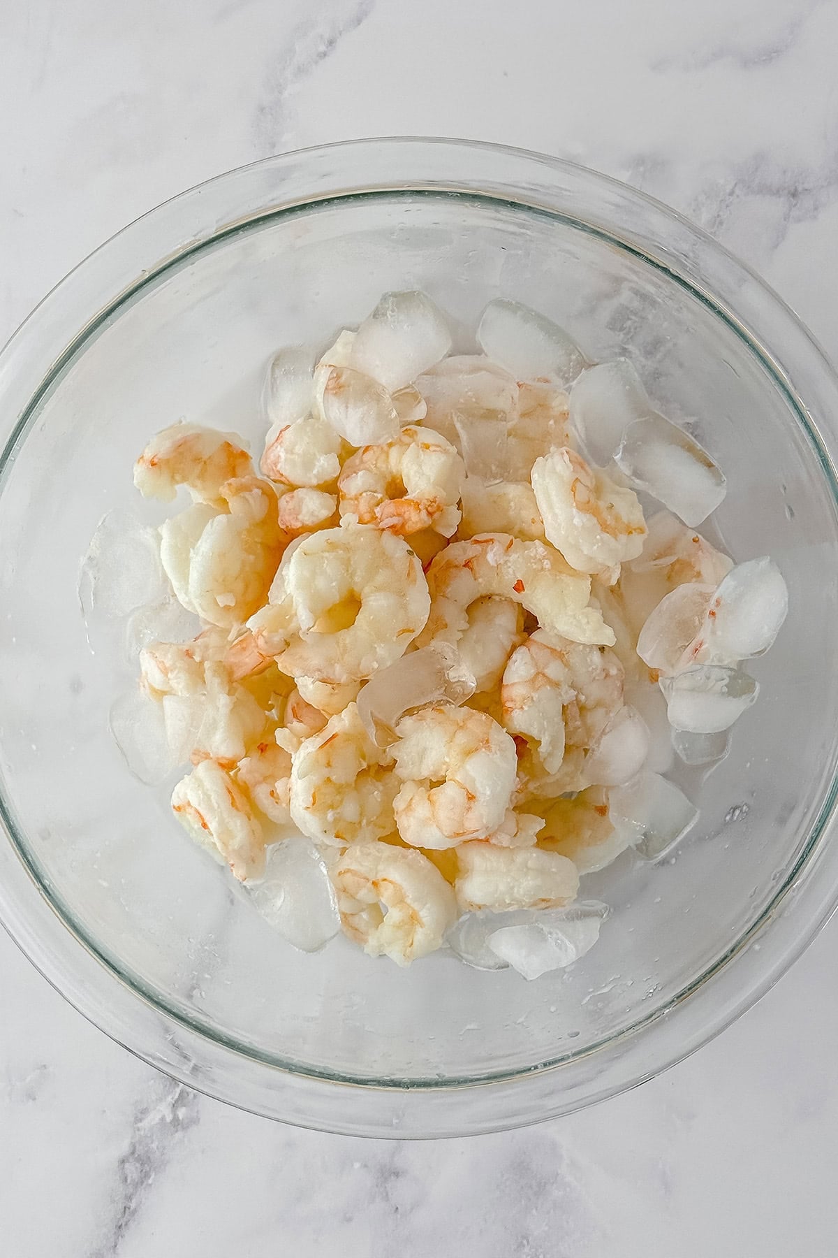 Cooked shrimp in a glass bowl surrounded by ice cubes on a white marble surface, perfect for creating classic New England Style Shrimp Rolls.