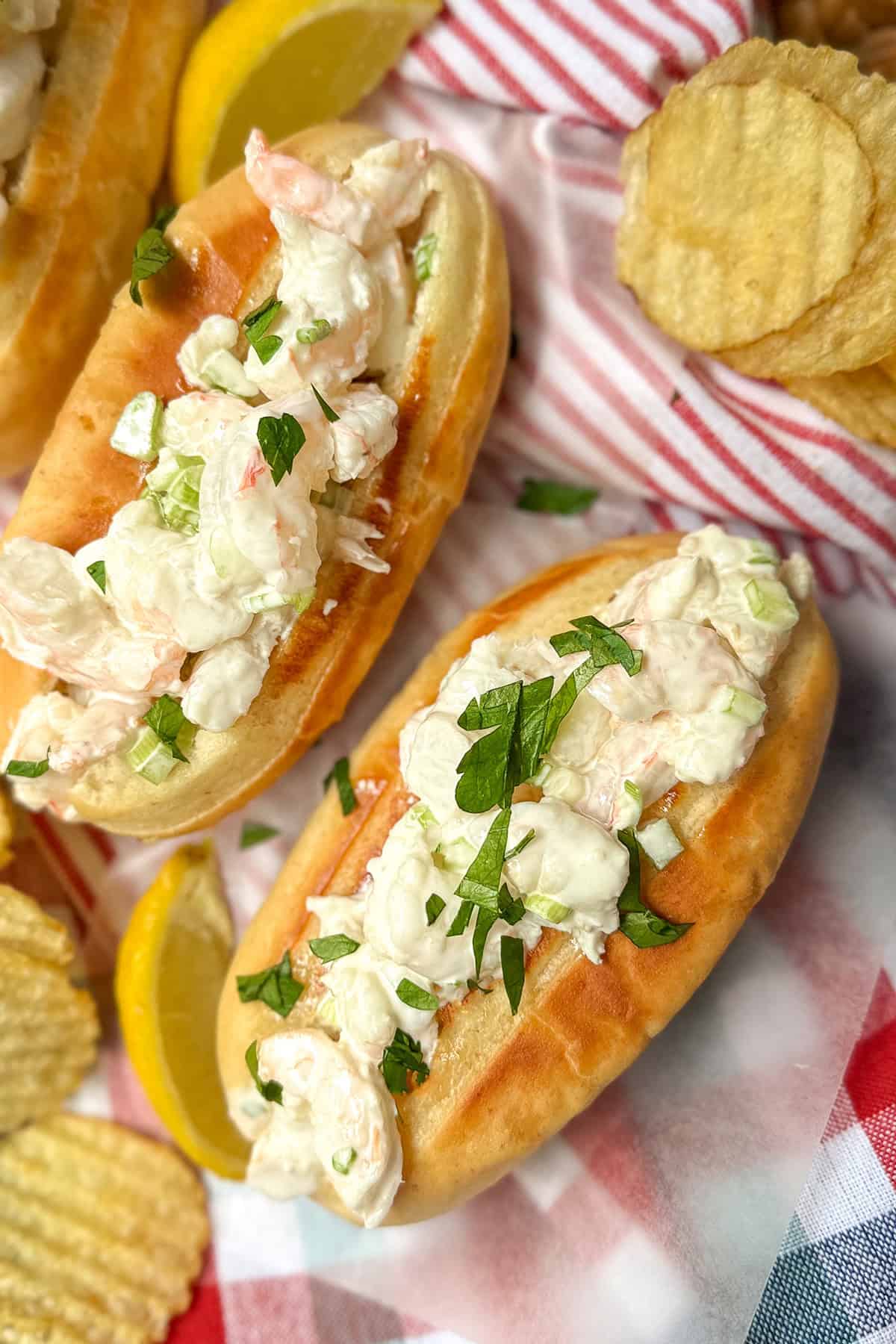 Two New England shrimp rolls garnished with fresh herbs on a bun, served with lemon wedges and potato chips atop a checkered cloth.