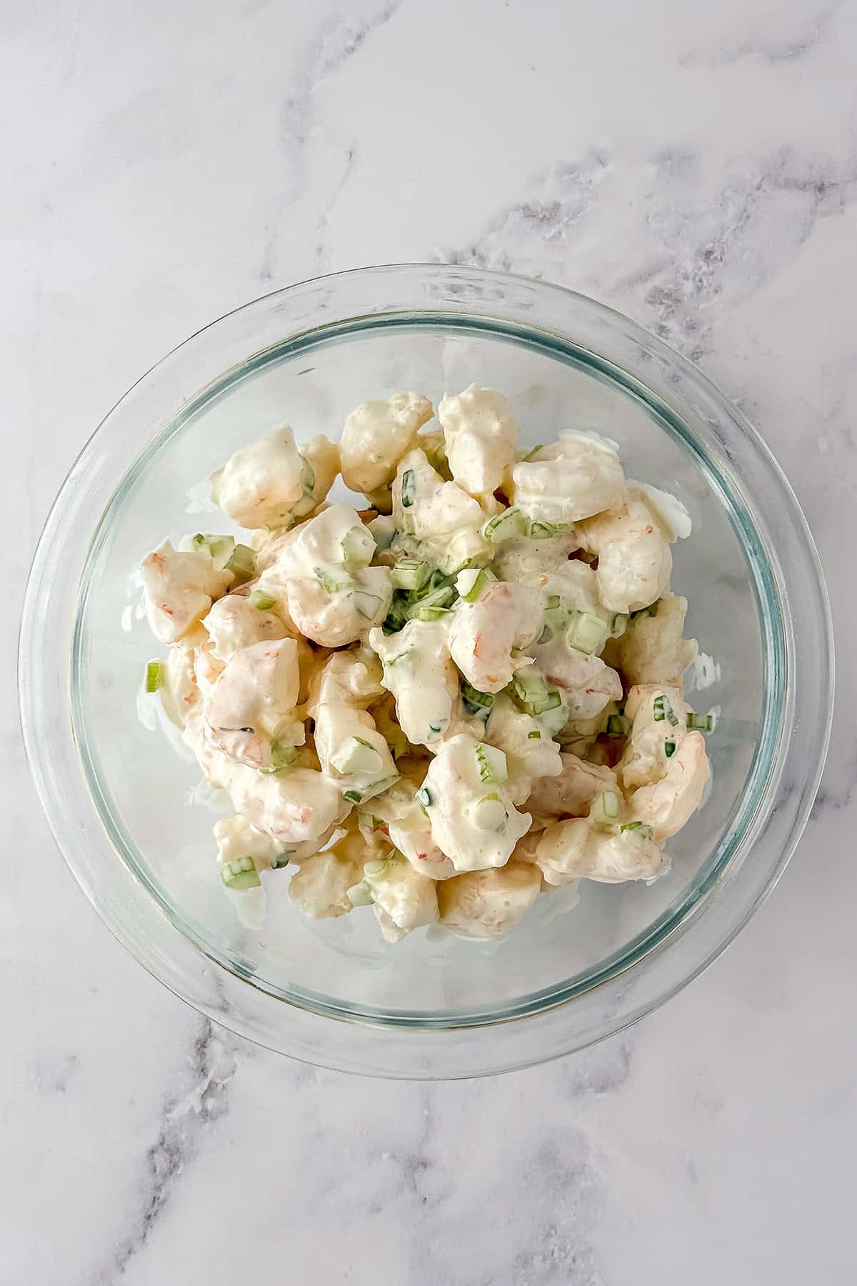 A glass bowl filled with a creamy shrimp and celery salad mixture-perfect for making classic New England Shrimp Rolls-sits on a white marble surface.