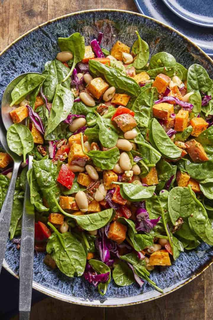 A large plate of spinach salad with white beans, roasted sweet potatoes, red bell peppers, and purple cabbage-perfect for May One Pot Dinners-served with metal utensils on a wooden table.