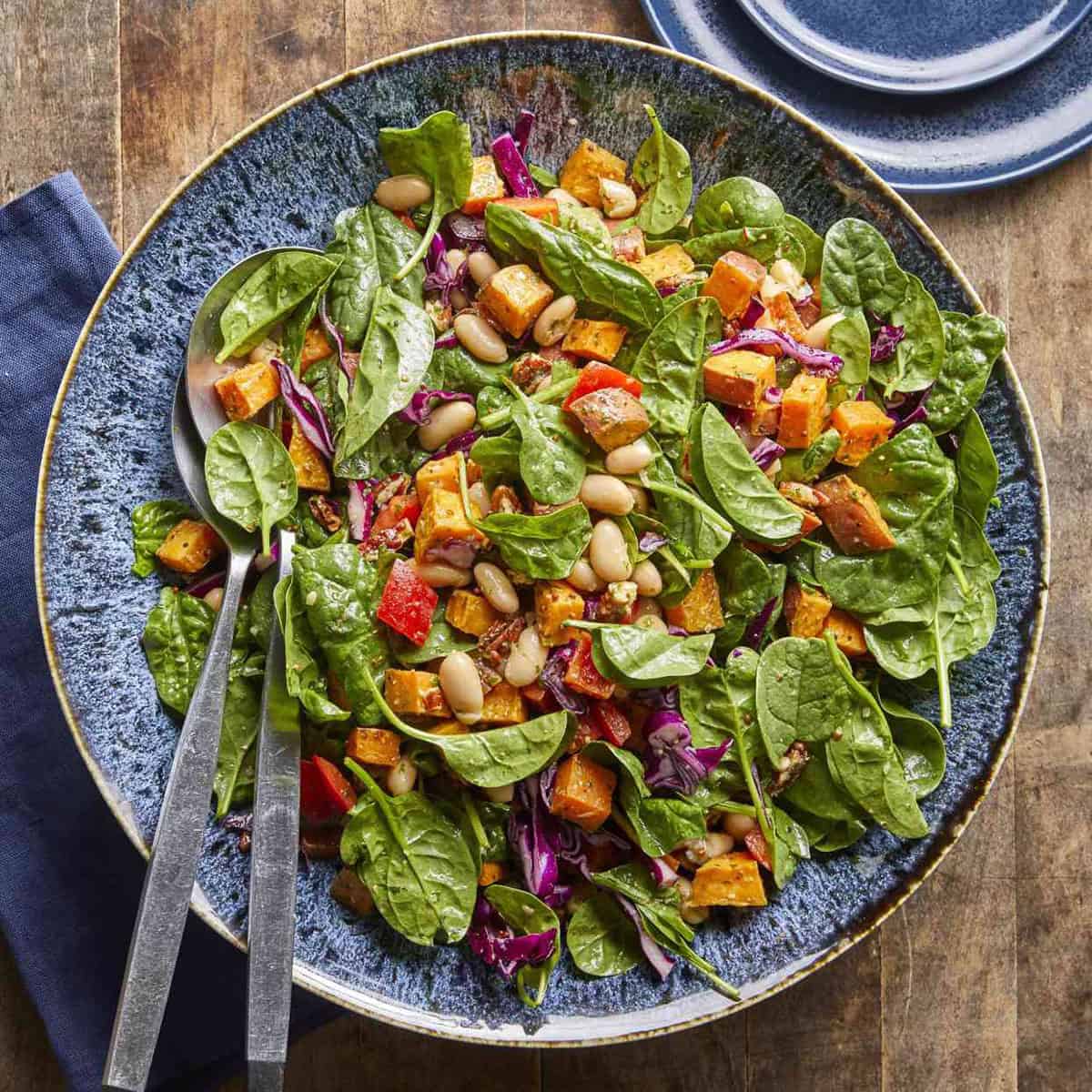 A large plate of spinach salad with white beans, roasted sweet potatoes, red bell peppers, and purple cabbage-perfect for May One Pot Dinners-served with metal utensils on a wooden table.