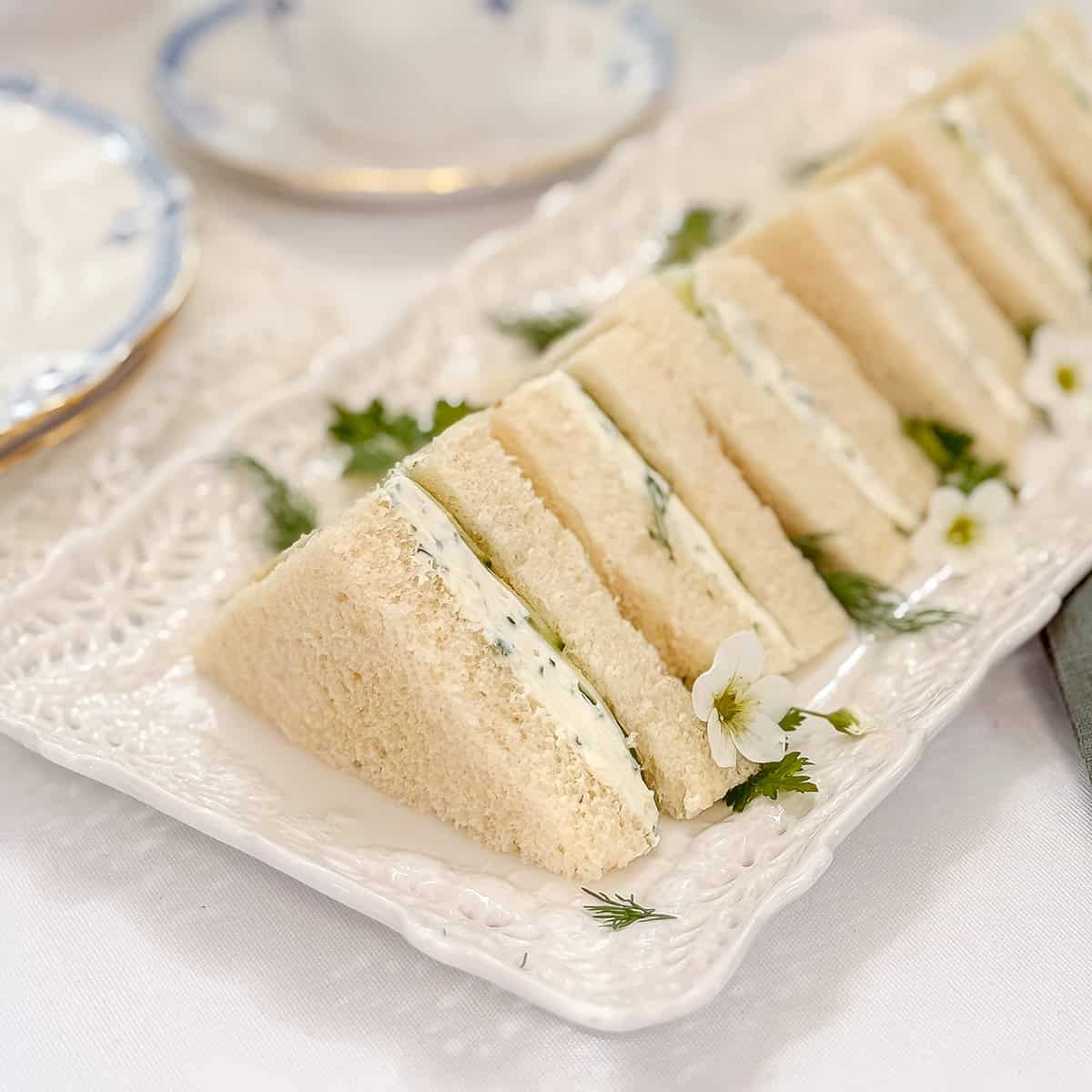 A plate of triangle-cut tea sandwiches with a creamy herb filling, perfect for Afternoon Tea, garnished with fresh herbs and small white flowers on a white decorative platter-a delightful treat for a Rare Day in June.