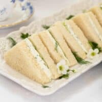 Rectangular plate with several triangular Benedictine Tea Sandwiches, filled with cream and herbs, garnished with small white flowers and dill, on a white tablecloth.