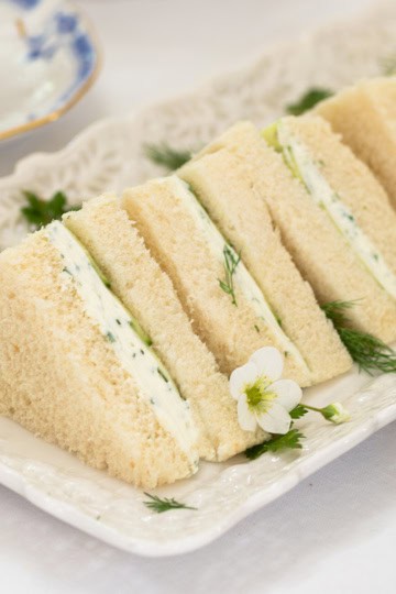 Rectangular plate with several triangular Benedictine Tea Sandwiches, filled with cream and herbs, garnished with small white flowers and dill, on a white tablecloth.