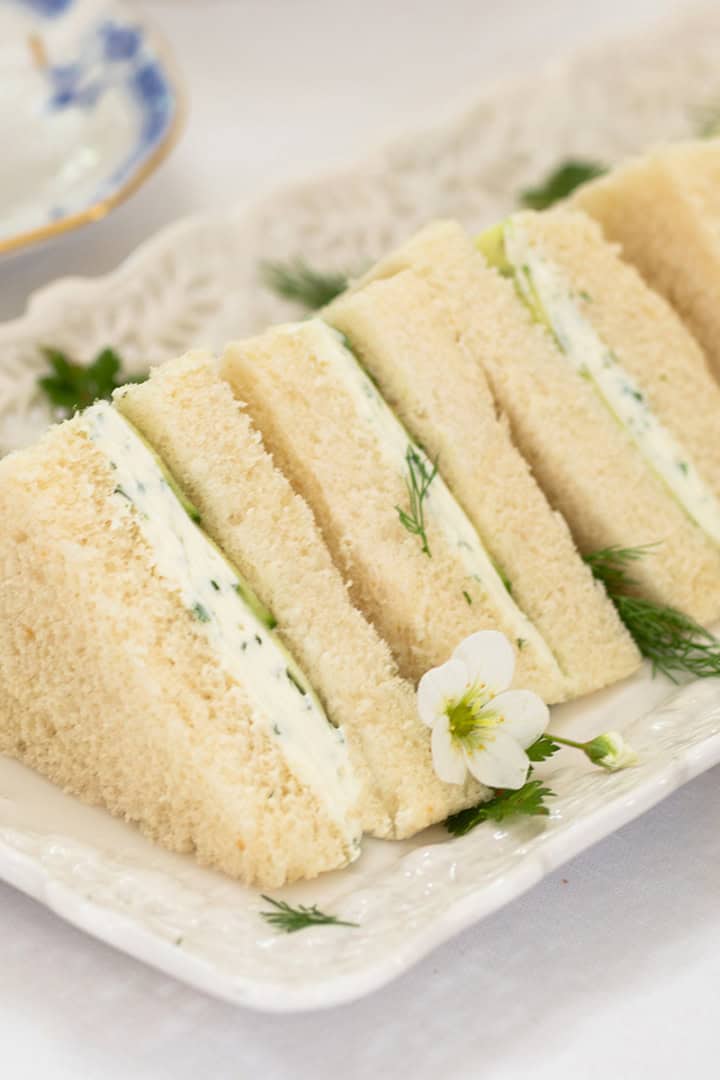 Rectangular plate with several triangular Benedictine Tea Sandwiches, filled with cream and herbs, garnished with small white flowers and dill, on a white tablecloth.