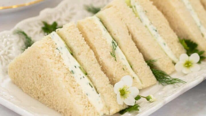 Rectangular plate with several triangular Benedictine Tea Sandwiches, filled with cream and herbs, garnished with small white flowers and dill, on a white tablecloth.