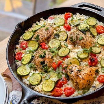 A one pan skillet brimming with Tuscan chicken thighs, sliced zucchini, cherry tomatoes, rice, and herbs, all beautifully arranged on a wooden surface.