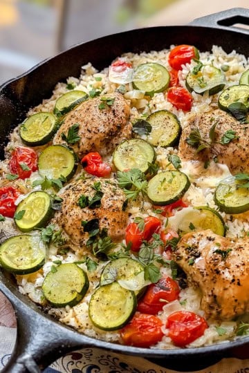 A one pan skillet brimming with Tuscan chicken thighs, sliced zucchini, cherry tomatoes, rice, and herbs, all beautifully arranged on a wooden surface.