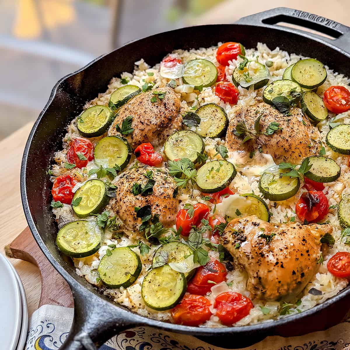 A one pan skillet brimming with Tuscan chicken thighs, sliced zucchini, cherry tomatoes, rice, and herbs, all beautifully arranged on a wooden surface.