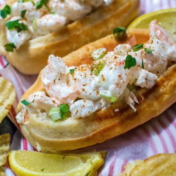 A classic New England shrimp roll on a toasted bun, garnished with chopped herbs and served with lemon wedges and crispy potato chips, delivers the ultimate seafood experience.
