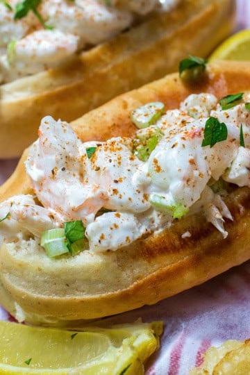A classic New England shrimp roll on a toasted bun, garnished with chopped herbs and served with lemon wedges and crispy potato chips, delivers the ultimate seafood experience.