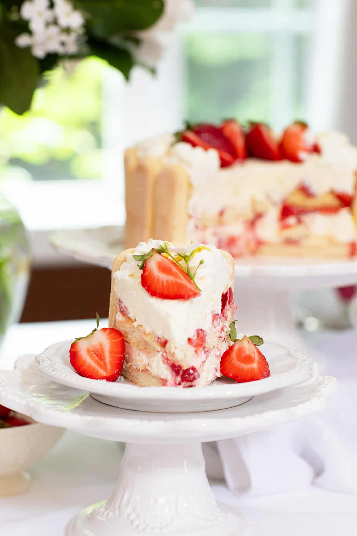 A slice of Strawberry Lady Charlotte Cake with whipped cream and fresh strawberries sits on a white cake stand, with the rest of the cake in the background.