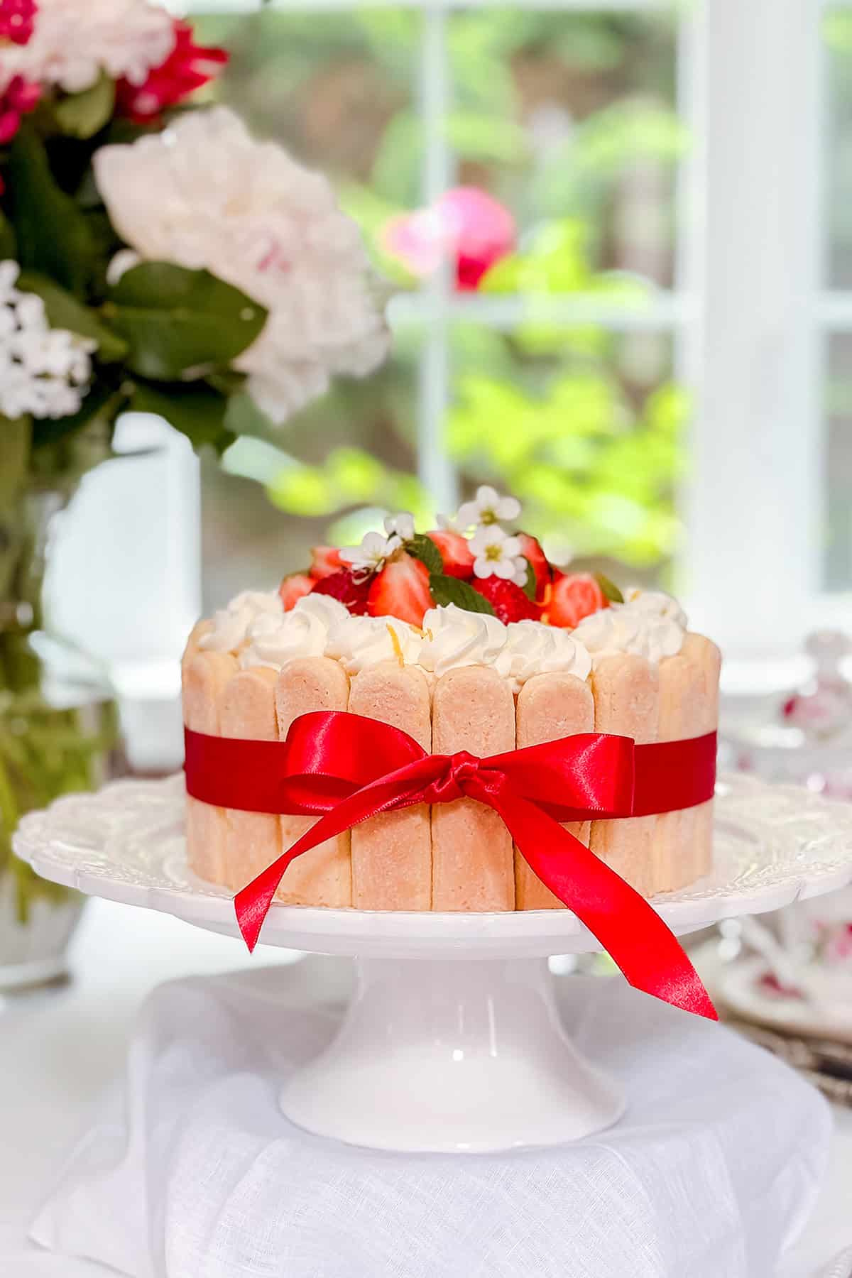 A decorated lady charlotte cake with ladyfingers, topped with strawberries and cream, wrapped with a red ribbon, displayed on a white cake stand.