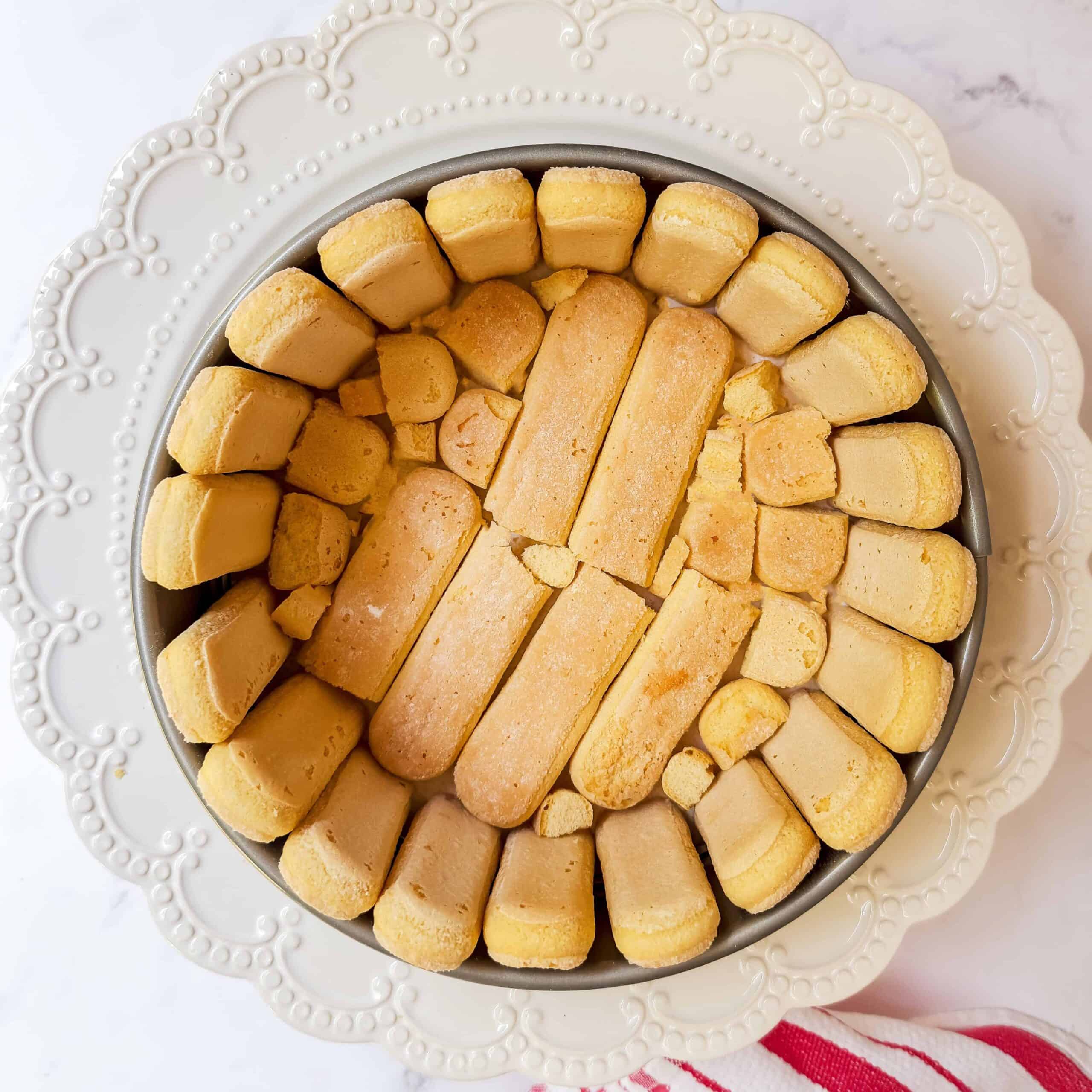 A round lady charlotte cake pan lined with ladyfinger biscuits, arranged neatly in concentric circles, sits on a white decorative plate next to a red and white striped cloth.