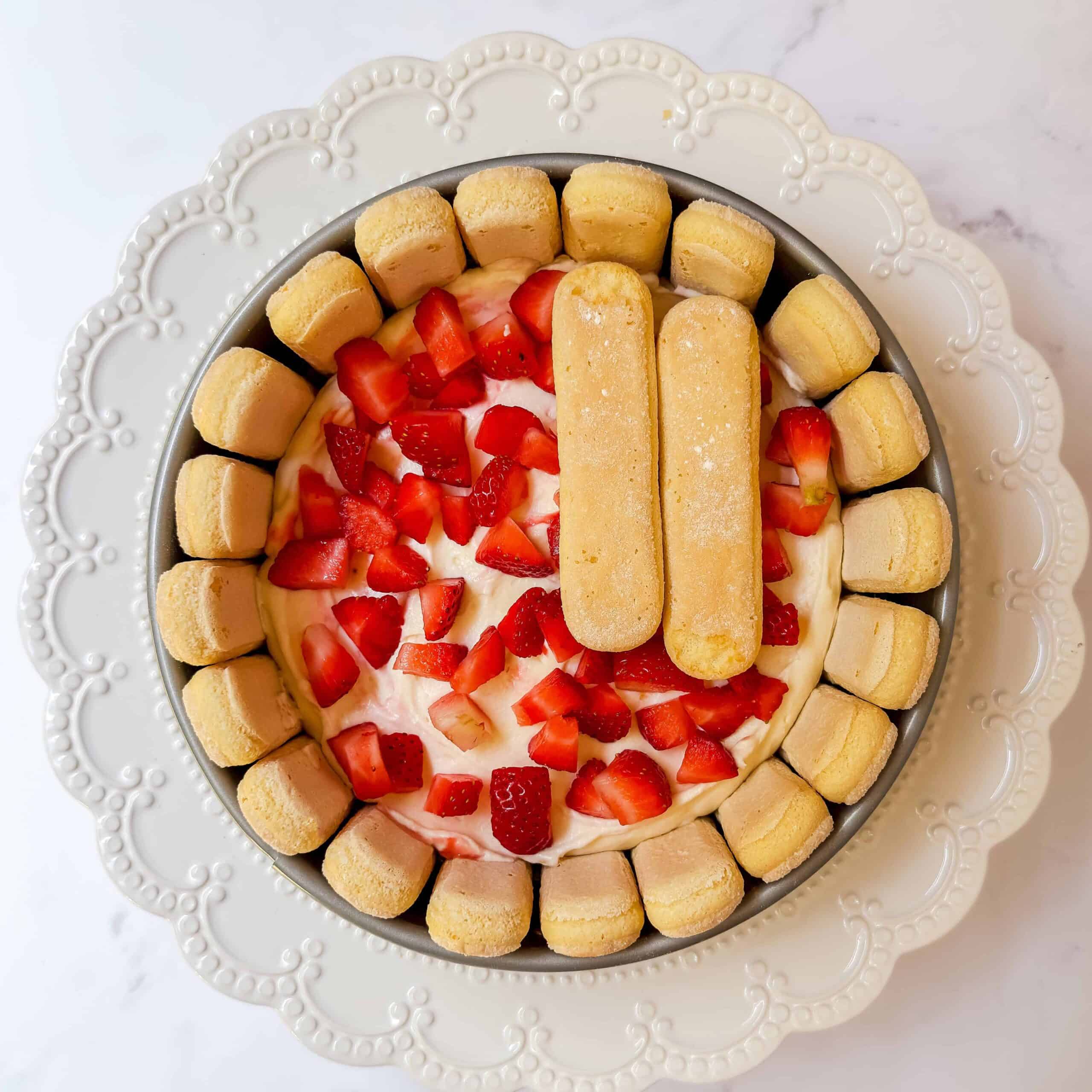 A round lady charlotte cake topped with chopped strawberries and two ladyfinger biscuits, surrounded by ladyfingers, on a white decorative plate.