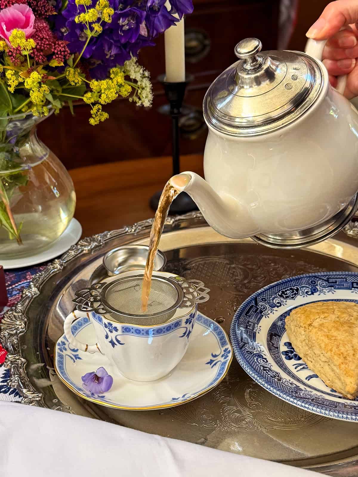 A hand pours Liberty TEa from a teapot through a strainer into a cup on a tray, with a scone on a plate and a vase of flowers nearby.