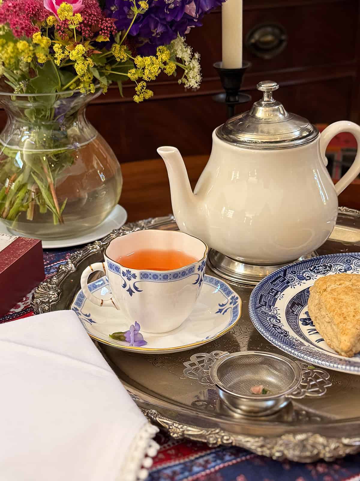 A white teapot, a cup of Liberty Tea, a plate with a scone, a napkin, and a vase of flowers are arranged on a silver tray.