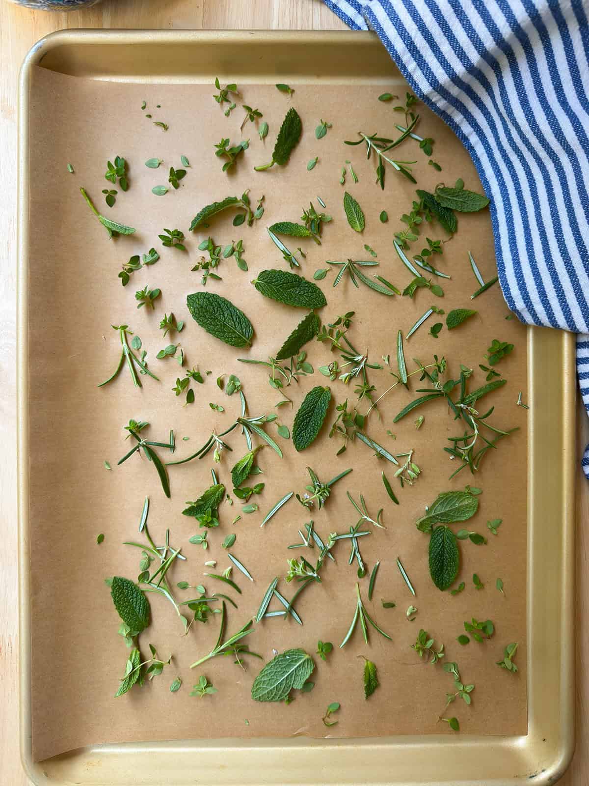 A baking sheet lined with parchment paper holds scattered fresh herbs, perfect for a Liberty Tea recipe, including mint, rosemary, and oregano leaves. A blue and white striped cloth is draped in the top right corner.