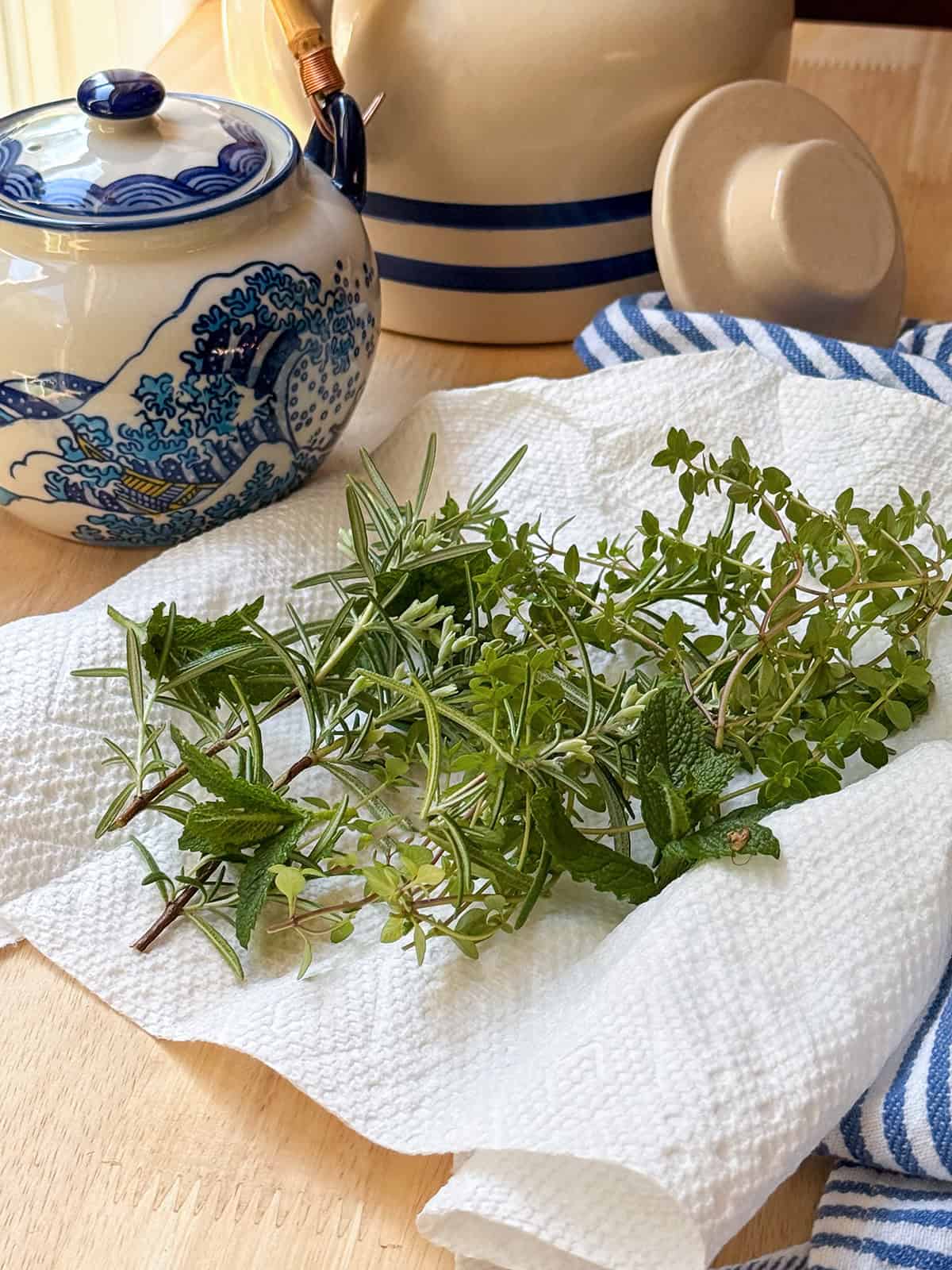 A bundle of fresh herbs, perfect for a Liberty Tea recipe, rests on a paper towel beside a ceramic teapot and a jar with blue patterns, all arranged on a wooden table with a striped cloth.