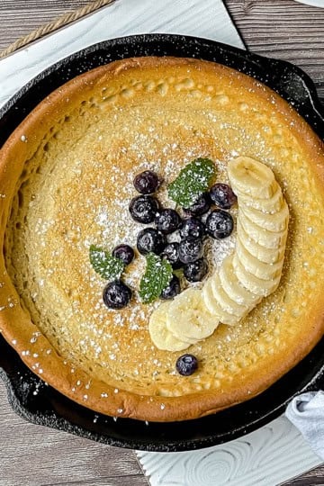 A perfect Father's Day Breakfast: a baked pancake in a cast iron skillet, topped with sliced bananas, blueberries, fresh mint leaves, and a dusting of powdered sugar.