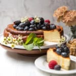 Basque Cheesecake topped with blackberries, blueberries, and raspberries, garnished with mint and flowers, on a cake stand with a slice served on a plate; flowers in the background.