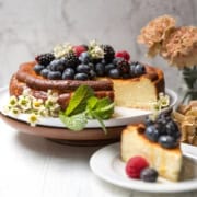 Basque Cheesecake topped with blackberries, blueberries, and raspberries, garnished with mint and flowers, on a cake stand with a slice served on a plate; flowers in the background.