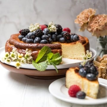 Basque Cheesecake topped with blackberries, blueberries, and raspberries, garnished with mint and flowers, on a cake stand with a slice served on a plate; flowers in the background.