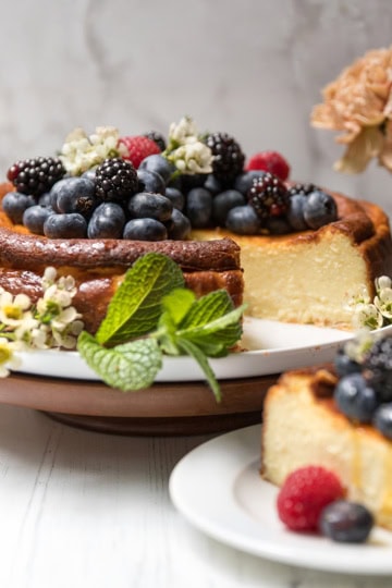 Basque Cheesecake topped with blackberries, blueberries, and raspberries, garnished with mint and flowers, on a cake stand with a slice served on a plate; flowers in the background.