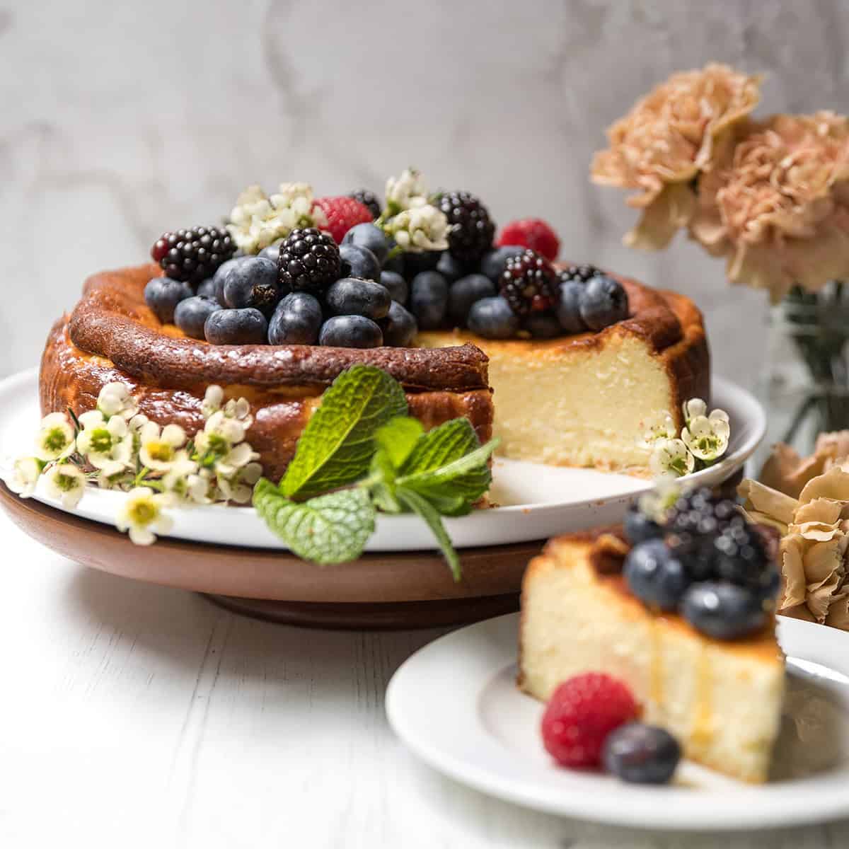 Basque Cheesecake topped with blackberries, blueberries, and raspberries, garnished with mint and flowers, on a cake stand with a slice served on a plate; flowers in the background.