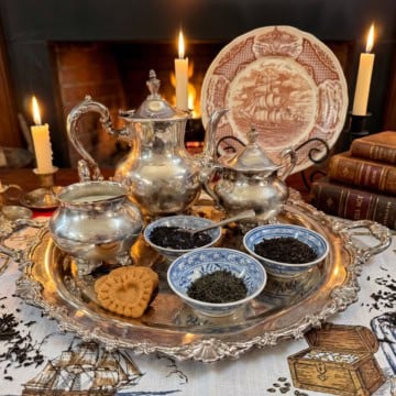Close-up of loose leaf teas displayed in blue and white Chinese tea bowls on an ornate silver tray, with a heart-shaped colonial shortbread and silver tea service, fireplace glowing warmly behind.
