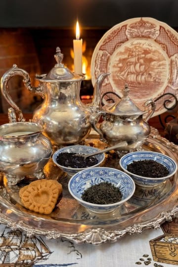 Close-up of loose leaf teas displayed in blue and white Chinese tea bowls on an ornate silver tray, with a heart-shaped colonial shortbread and silver tea service, fireplace glowing warmly behind.