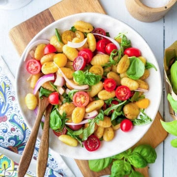 A plate of gnocchi salad with cherry tomatoes, arugula, spinach, red onion, and basil, served with wooden utensils on a light table.