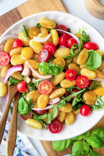 A plate of gnocchi salad with cherry tomatoes, arugula, spinach, red onion, and basil, served with wooden utensils on a light table.