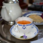 A blue and white porcelain teacup filled with tea, inspired by a classic Liberty Tea recipe, sits on a matching saucer beside a teapot and a small purple flower, all arranged on a metal tray.