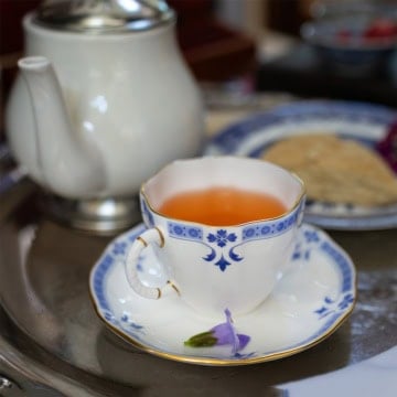 A blue and white porcelain teacup filled with tea, inspired by a classic Liberty Tea recipe, sits on a matching saucer beside a teapot and a small purple flower, all arranged on a metal tray.