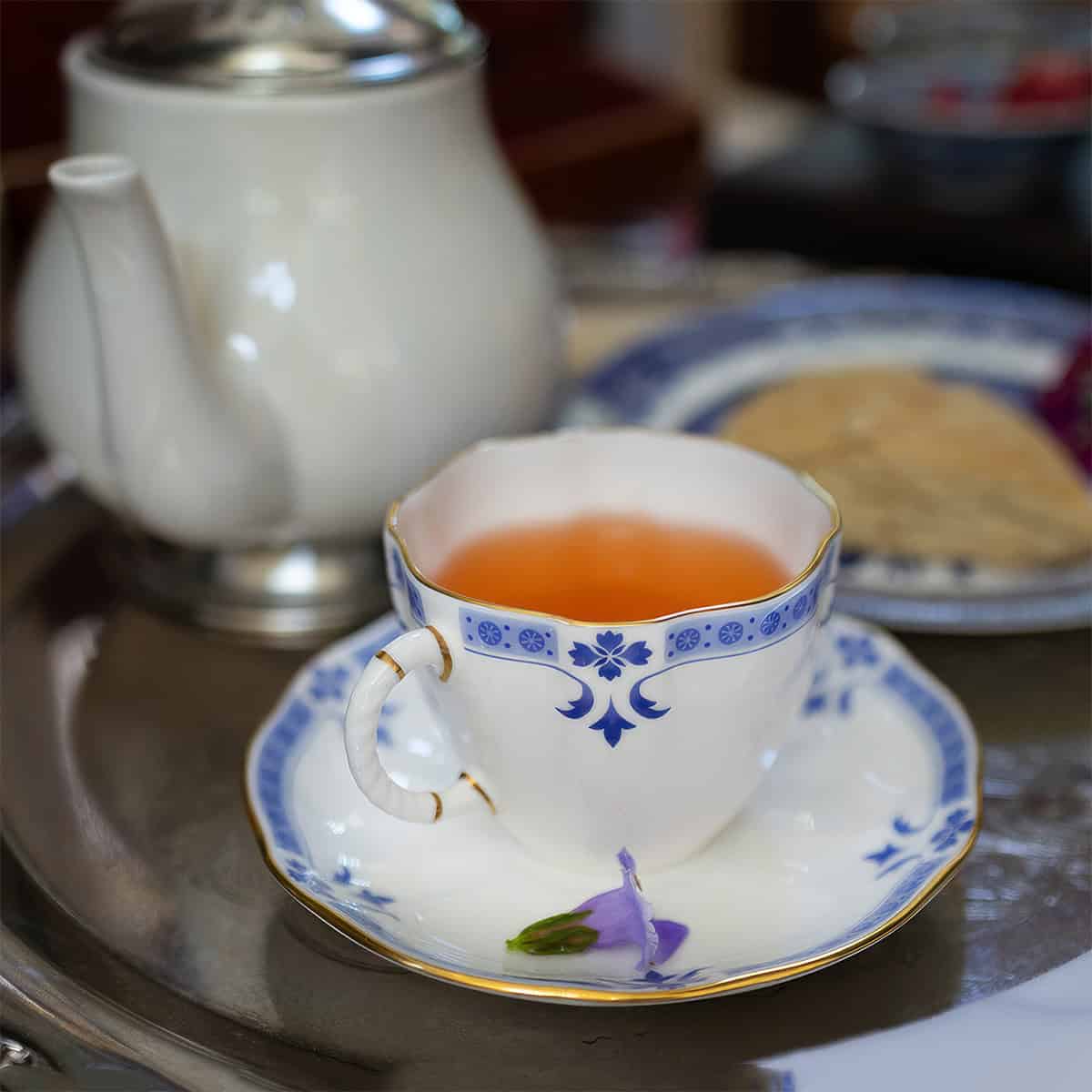 A blue and white porcelain teacup filled with tea, inspired by a classic Liberty Tea recipe, sits on a matching saucer beside a teapot and a small purple flower, all arranged on a metal tray.