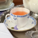 A cup of pink Liberty Tea in a white and blue teacup on a matching saucer, with a small purple flower resting beside it, placed elegantly on a silver tray.