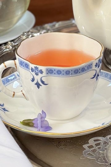 A cup of pink Liberty Tea in a white and blue teacup on a matching saucer, with a small purple flower resting beside it, placed elegantly on a silver tray.