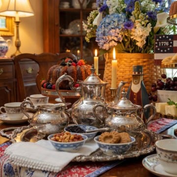 A silver tea set with cups, bowls of snacks, and a bundt cake arranged on a dining table, with candles, flowers, patriotic decor, and a hint of Revolutionary Tea spirit in the background.