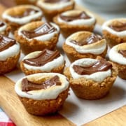 Bite-sized s'mores cups with toasted marshmallow and melted chocolate on a graham cracker base, arranged on a wooden board.