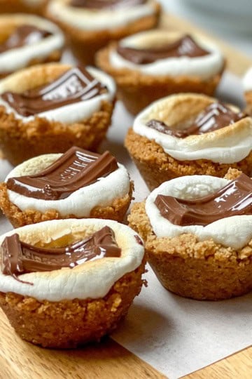 Bite-sized s'mores cups with toasted marshmallow and melted chocolate on a graham cracker base, arranged on a wooden board.